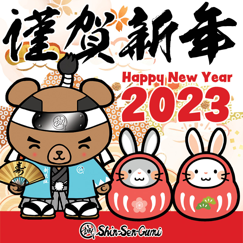 "Kinga Shin Nen" Kanji Black letters on the top, Happy New Year 2023 in Red letters in the middle, Smiling Shin-Sen-Bear has a golden Japanese folding fan on the right hand. Next to him, 2 rabbits wearing DARUMA costumes, Shin-Sen-Gumi logo on the bottom.